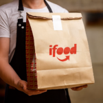 Entregador segurando sacola ifood em ambiente interno.