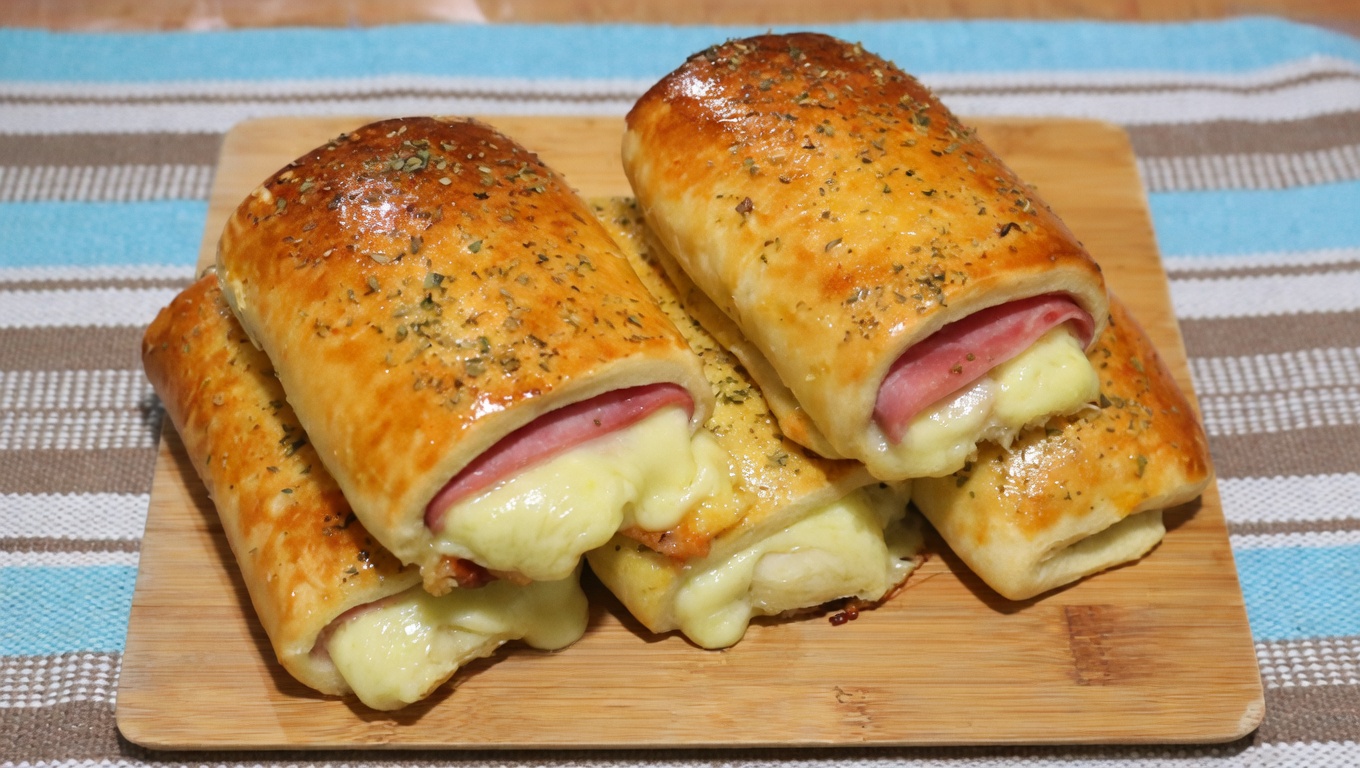 joelhos com massa dourada recheados com presunto e queijo derretido, polvilhados com ervas, empilhados sobre uma tábua de madeira, com queijo cremoso escorrendo.