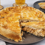 Empadão de frango caseiro dourado em prato cinza, com fatia sendo servida por espátula. A crosta crocante e dourada exibe pequenas esferas decorativas na borda, revelando recheio de frango desfiado úmido. Ao fundo, copos de guaraná com bolhas sobre descansos de madeira e toalha listrada em tons neutros completam o cenário iluminado naturalmente, com aparência realista e refinada.